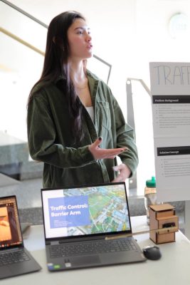 A student stands behind her laptop that has a screen reading "Traffic Control: Barrier Arm." She stands next to a posterboard about Traffic Control on Drillfield Drive.