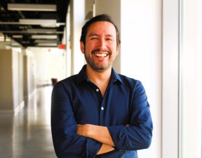 Ernesto is smiling into the camera with his arms crossed while wearing a blue shirt 